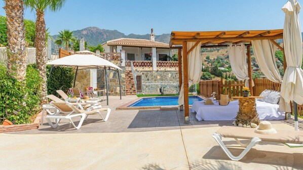 Uma área de piscina com espreguiçadeiras, uma pérgula com cortinas e vista para montanhas ao fundo.