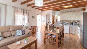 A cozy living room with a sofa, coffee table, and dining area. A kitchen with wooden cabinets and a refrigerator is visible in the background. - Villa Gamor Frigiliana by Ruralidays (Málaga)