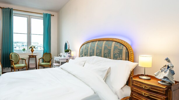 A hotel room with a large bed, a dresser with drawers, a nightstand with a lamp, a chair, a desk with a mirror, and a window with curtains. - luxury studio 5 (Hlavní mesto Praha)