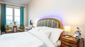 A hotel room with a large bed, a dresser with drawers, a nightstand with a lamp, a chair, a desk with a mirror, and a window with curtains.