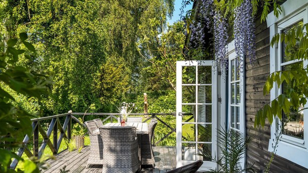 A wooden deck with wicker furniture and potted plants, overlooking a lush garden and a white building with a large window. - Stunning home in Sjællands Odde (Sjællands Odde)