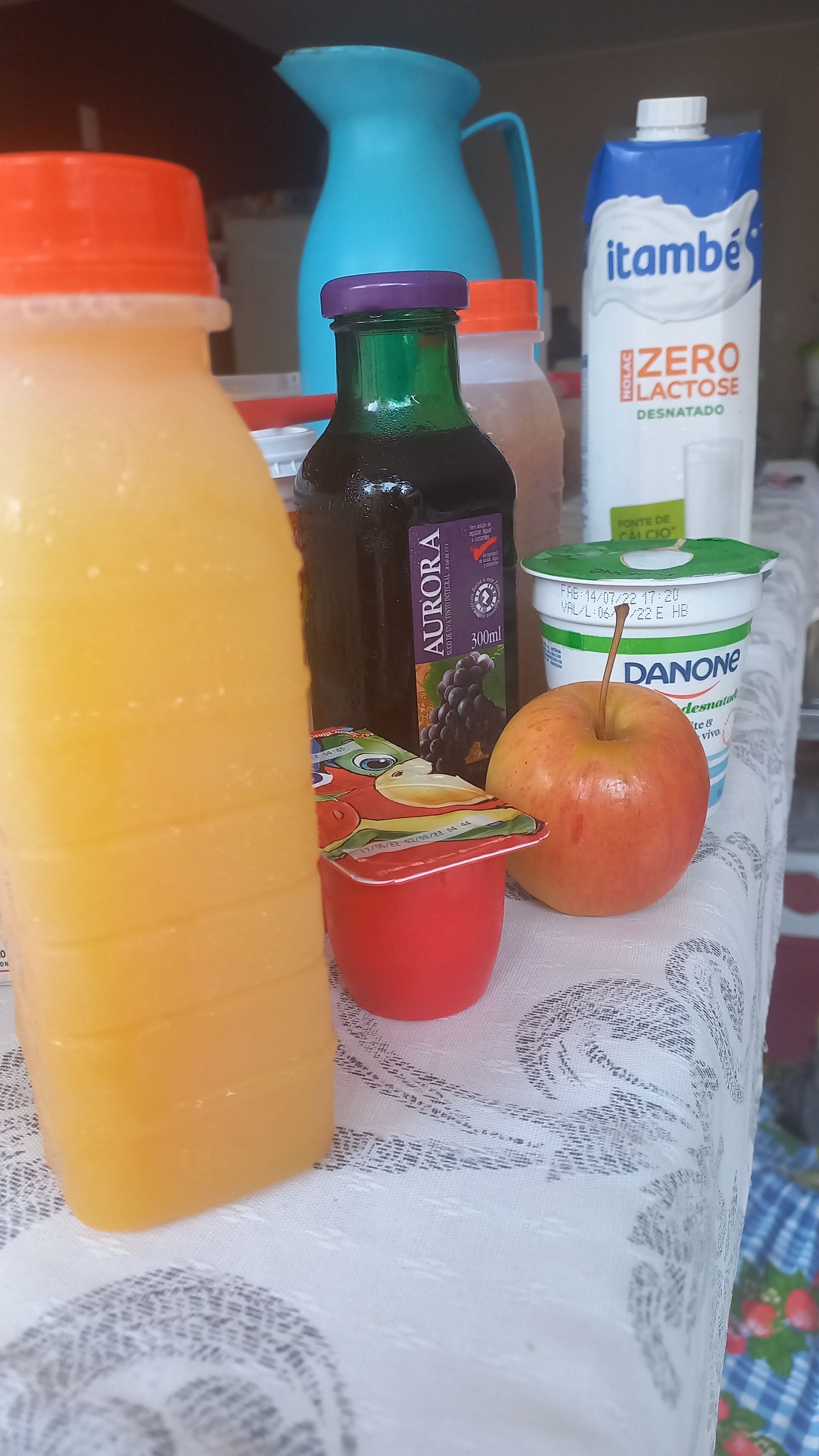 Une table sur laquelle se trouvent divers aliments, notamment une bouteille de jus d’orange, une bouteille contenant un liquide foncé, un emballage de yaourt Danone, un emballage de lait Itambe sans lactose, une tasse rouge et une pomme.