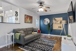 A modern living room with a gray sofa, a black and white rug, a ceiling fan, a mounted TV, and a wooden shelf with decorative items.
