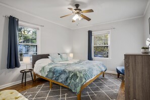 A bedroom with a bed, a ceiling fan, two windows with curtains, a bedside table, and a nightstand.