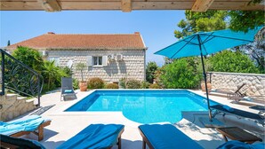 A pool area with lounge chairs and a blue umbrella, a stone house in the background, and a wooden pergola overhead. - Amazing home in Cavtat (Cavtat)