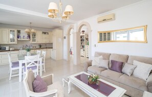 A spacious living area with a sofa, coffee table, and dining set. There is a kitchen in the background with white cabinets and a chandelier.