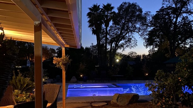 An outdoor swimming pool area with a covered patio, seating, and surrounding vegetation.
