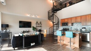 Un apartamento moderno con cocina, comedor y sala de estar. Cuenta con un sofá negro, un televisor, una escalera y una cocina con armarios de madera y un taburete azul.