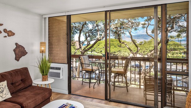 Un salon moderne avec un canapé marron, une table basse blanche et une grande porte-fenêtre coulissante donnant sur une terrasse extérieure, d’où l’on peut admirer des arbres et un paysage lointain.
