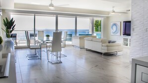 A modern living room with a large window overlooking the sea, a white sofa, a dining area with chairs, and a TV mounted on the wall. - VillaPlayaEncantada-Pools&BeachAccess-Rosarito (Rosarito)