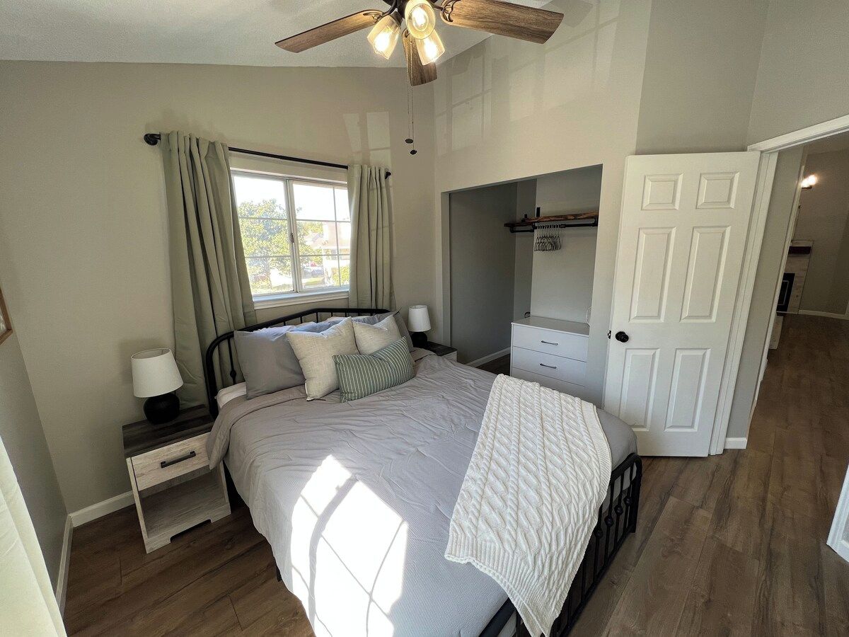 A bedroom with a bed, nightstands, a window with curtains, a ceiling fan, and a door leading to another room.