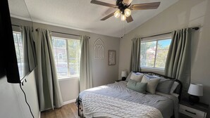 Un dormitorio con cama, mesitas de noche, ventilador de techo y ventana con cortinas.
