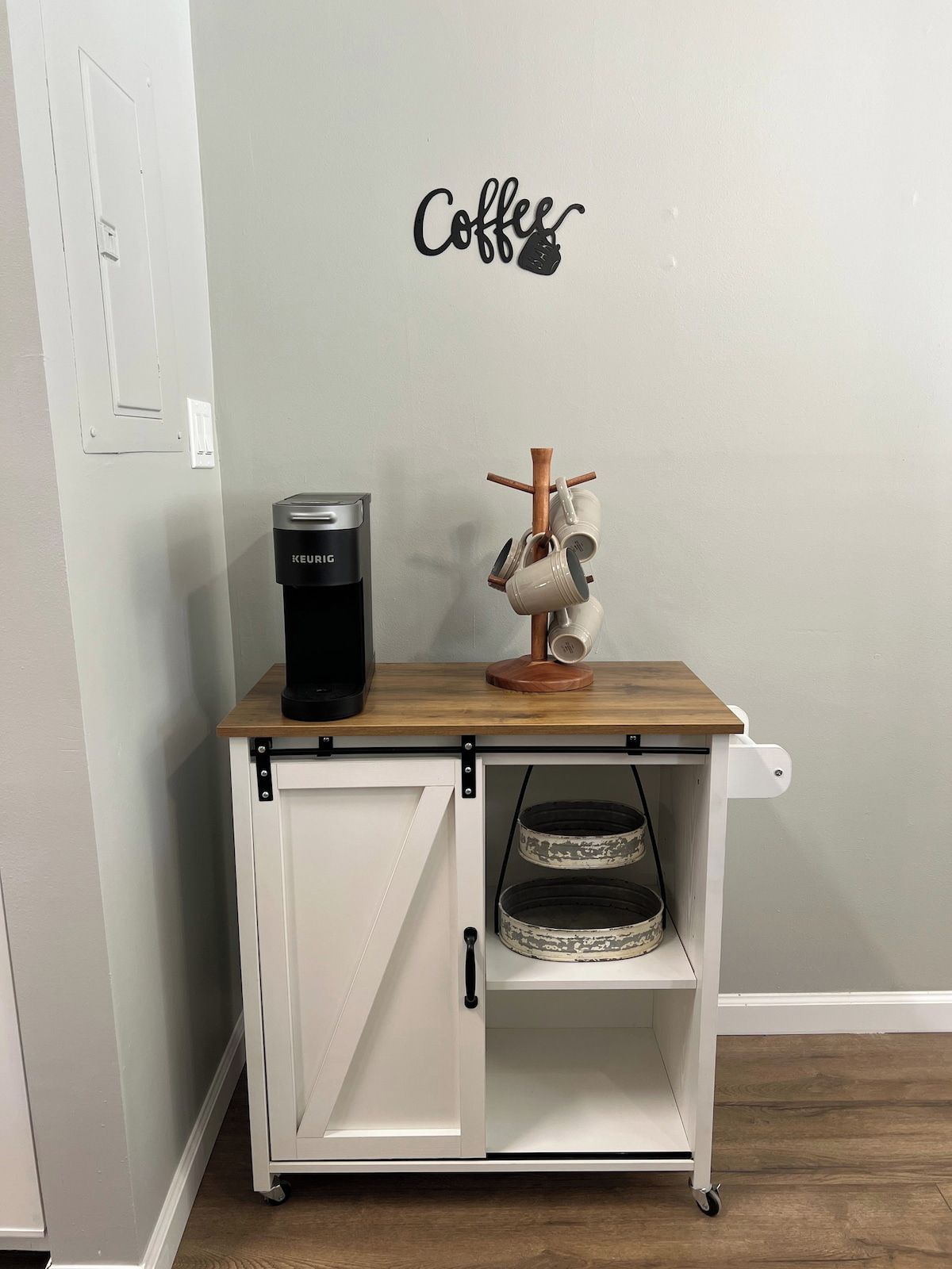 A white cabinet with a coffee maker and mug holder, a shelf with a patterned basket, and a wall sign that says 'Coffee'.