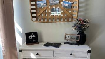 A white desk with a basket of printed photos, a welcome sign, a relaxation sign, and a small plant on a shelf.