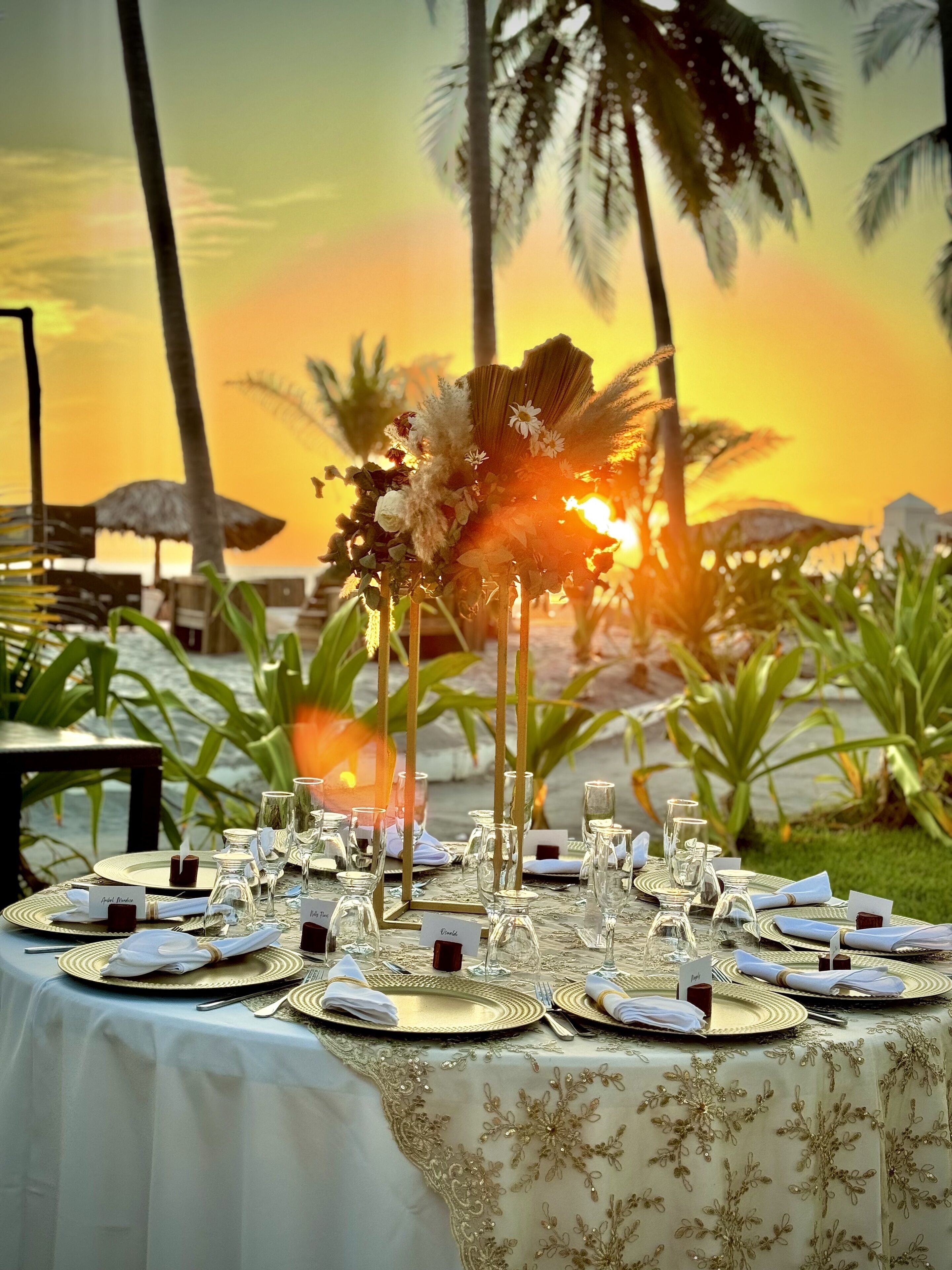 A table set for a meal with a sunset view, palm trees, and a thatched umbrella in the background.