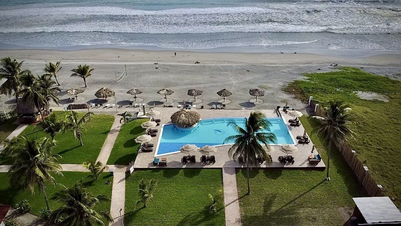 An aerial view of a beachside resort with a central swimming pool, lounge areas, and palm trees.