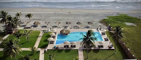 Vista aérea de un resort frente a la playa, con una piscina central, zonas de descanso y palmeras.