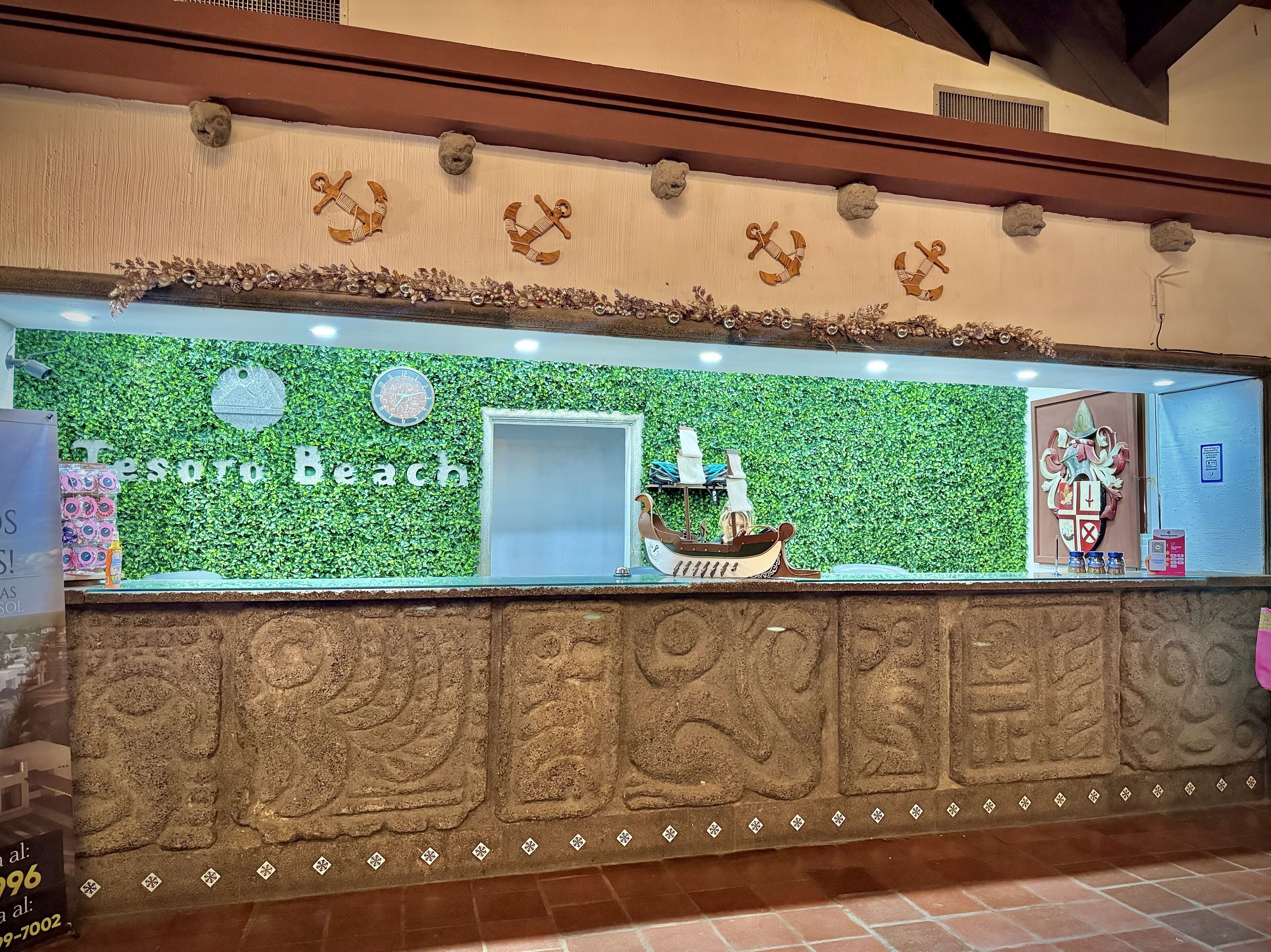 En reception på 'Tesoro Beach' med grøn væg, dekorative ankre, en skibsmodel og et ur.