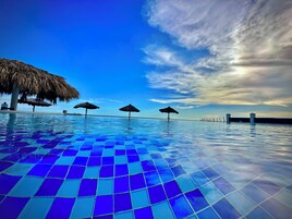 A swimming pool with a tiled floor and thatched umbrellas.