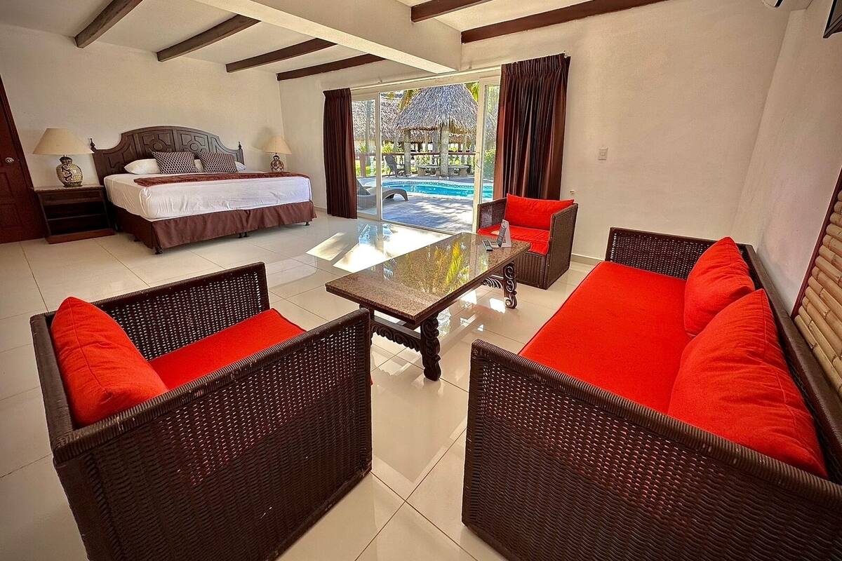 Ein Hotelzimmer mit Bett, Esstisch, Korbmöbeln und Blick auf einen Swimmingpool.