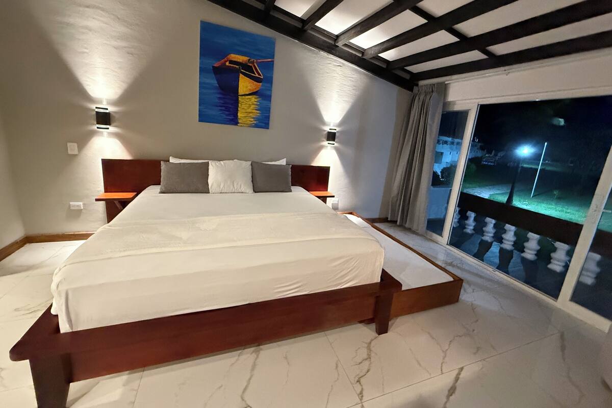 Ein geräumiges Schlafzimmer mit einem großen Bett, einem Holzkopfende, zwei Nachttischlampen, einem Gemälde über dem Bett und einem Balkon mit Blick ins Dunkel.