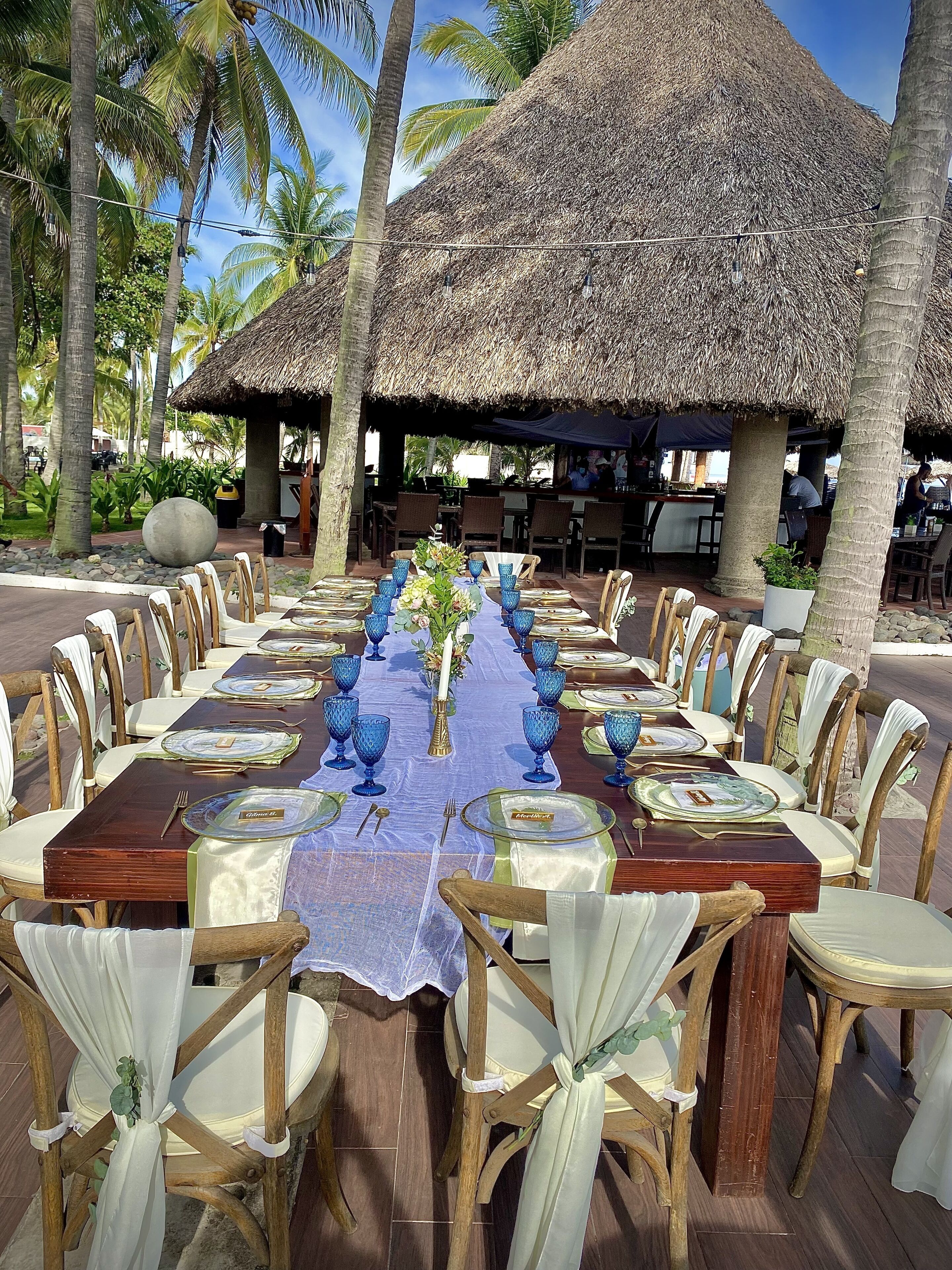 A long dining table set for a meal, surrounded by chairs, with a thatched roof structure in the background.