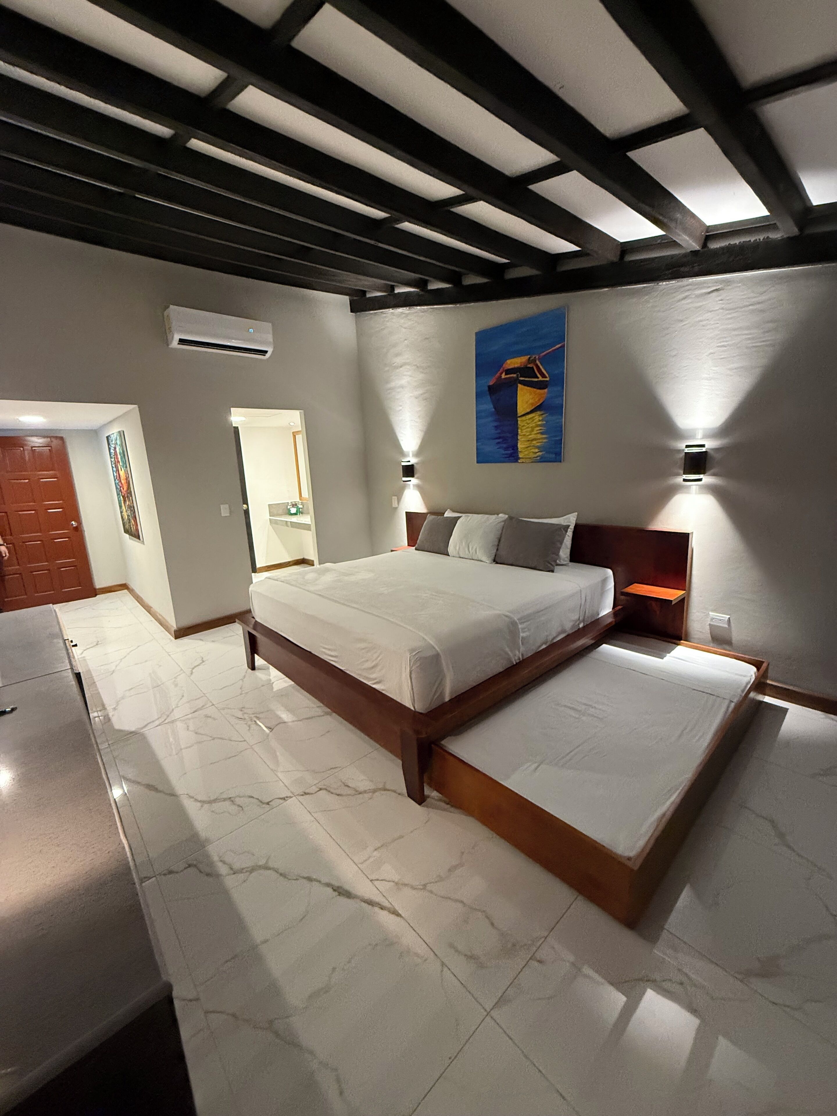 A bedroom with a large bed, a wooden headboard, and a nightstand with a lamp. There is a door and a painting on the wall.
