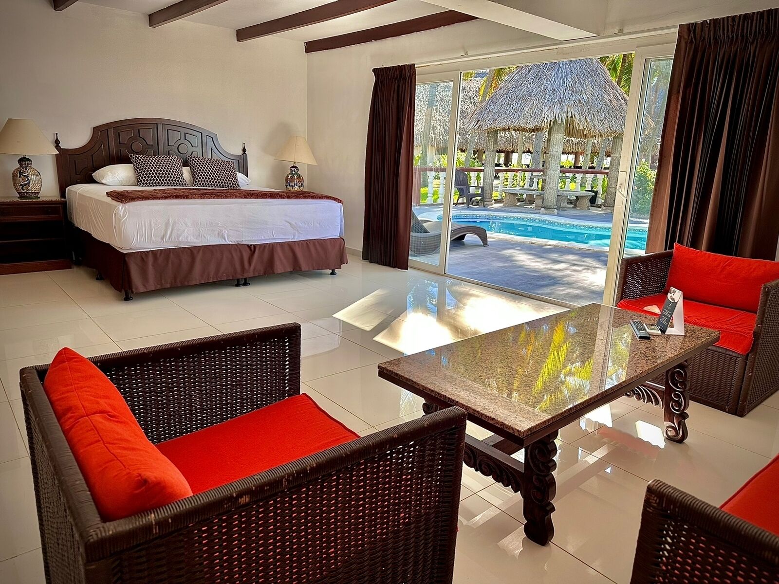 A hotel room with a bed, two wicker chairs with red cushions, a glass-top table, and a view of an outdoor pool area.