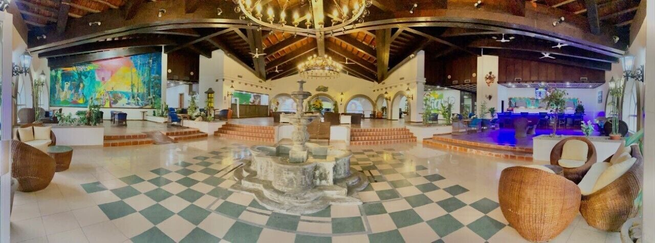 A spacious lobby with a checkered floor, a central fountain, wicker furniture, and a large mural on the wall.