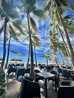 A beachside café with outdoor seating, palm trees, and a view of the ocean.