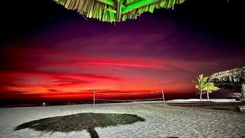 Een strandvolleybalveld met een net, palmbomen en een overkapping met rieten dak.
