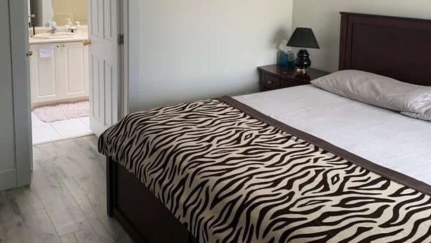 Un dormitorio con una colcha de estampado de cebra, un ventilador de techo, una lámpara de noche y vistas a un baño por una puerta abierta.