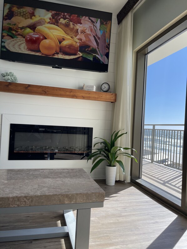 A modern living room with a flat-screen TV, a fireplace, a dining table, and a balcony with a view of the ocean.