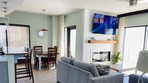 Ein modernes Wohnzimmer mit einem grauen Sofa, einem Holzesstisch und Stühlen. Ein Flachbildfernseher ist an der Wand befestigt, und ein Kamin ist sichtbar.