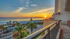 Ein Balkon mit Blick auf den Sonnenuntergang über dem Meer, Palmen und geparkte Autos.