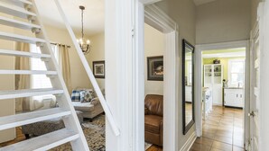 Un escalier mène à un salon agrémenté d’un lustre, d’un canapé en cuir marron et d’un coin repas donnant sur une cuisine visible à travers une porte ouverte.