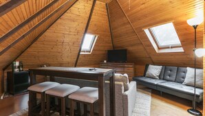 Une pièce mansardée chaleureuse avec un plafond en pente en bois, un canapé, une table avec des tabourets, une télévision et deux fenêtres de toit.