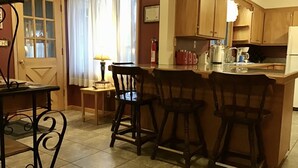 Een keuken met houten kastjes, een eettafel met stoelen, een klein bijzettafeltje met een lamp, en een raam met gordijnen.