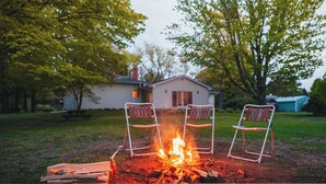 Een kampvuur met twee metalen stoelen in een achtertuin, met een huis op de achtergrond.