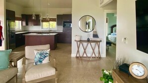 A modern kitchen with dark cabinets, a central island, and pendant lighting. There is a dining area with a sofa and a small table. A large mirror hangs on the wall, and a clock is visible on a wooden surface. - Havana Palms, 2 Bedroom Paradise (Milton Bight)