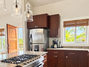 A kitchen with a stainless steel refrigerator, a gas stove, wooden cabinets, and a window with a view of greenery.