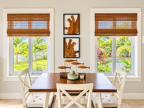 Un comedor con una mesa de madera, sillas blancas y dos ventanas con persianas de bambú.
