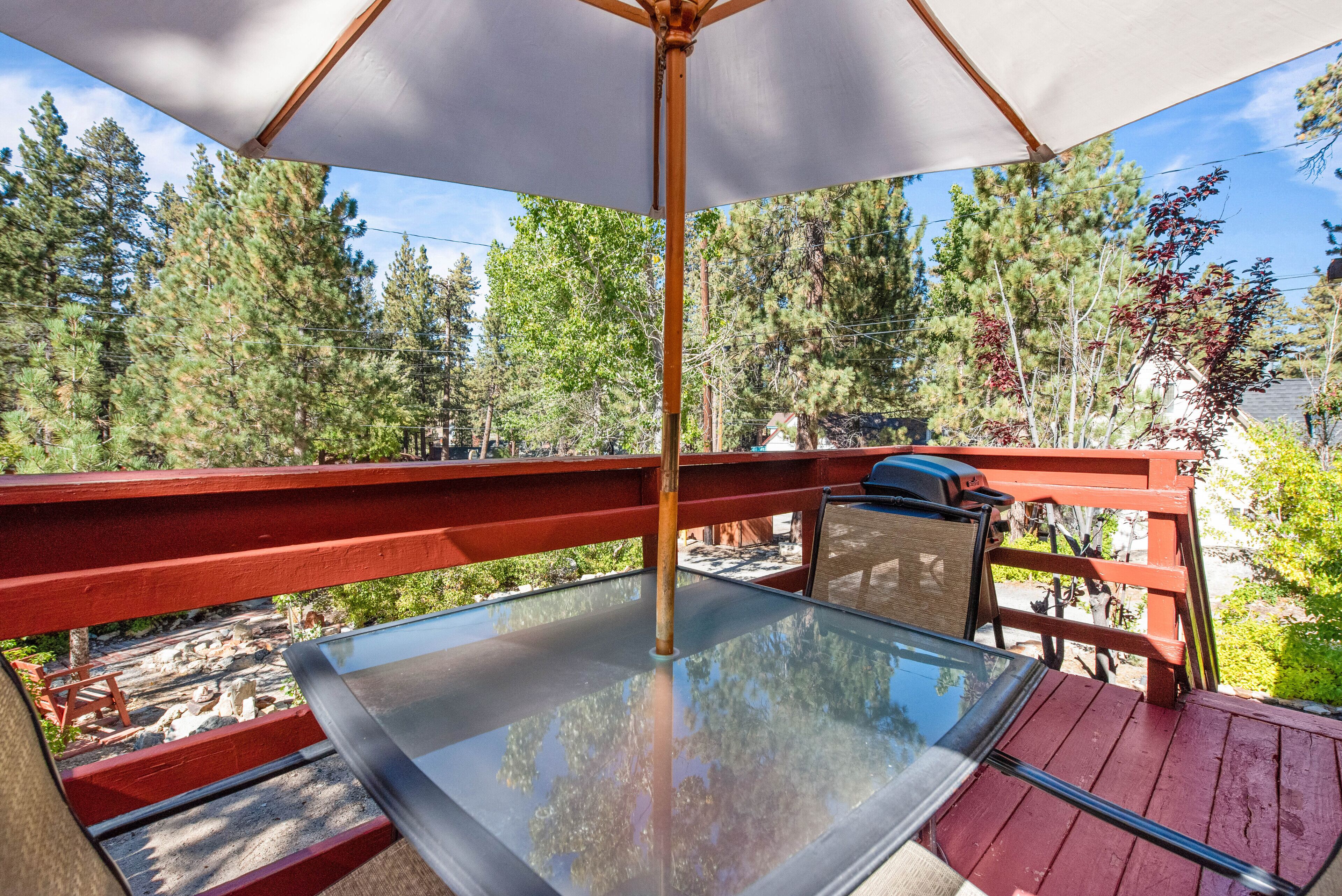 Une table en verre sous un parasol blanc, sur une terrasse rouge, avec vue sur les arbres et un barbecue.