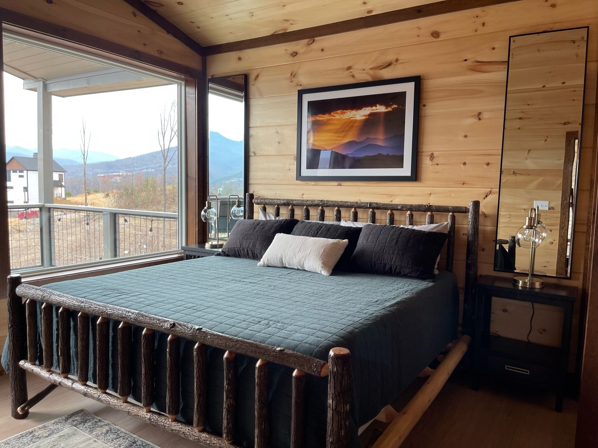 Un dormitorio con una cama grande, cabecera de madera, un cuadro enmarcado en la pared y vistas a las montañas desde la ventana.