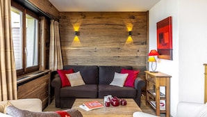Una sala de estar acogedora con una pared de madera, un sofá con cojines, una mesa de centro con un libro y tazas, y una lámpara sobre una mesita auxiliar.
