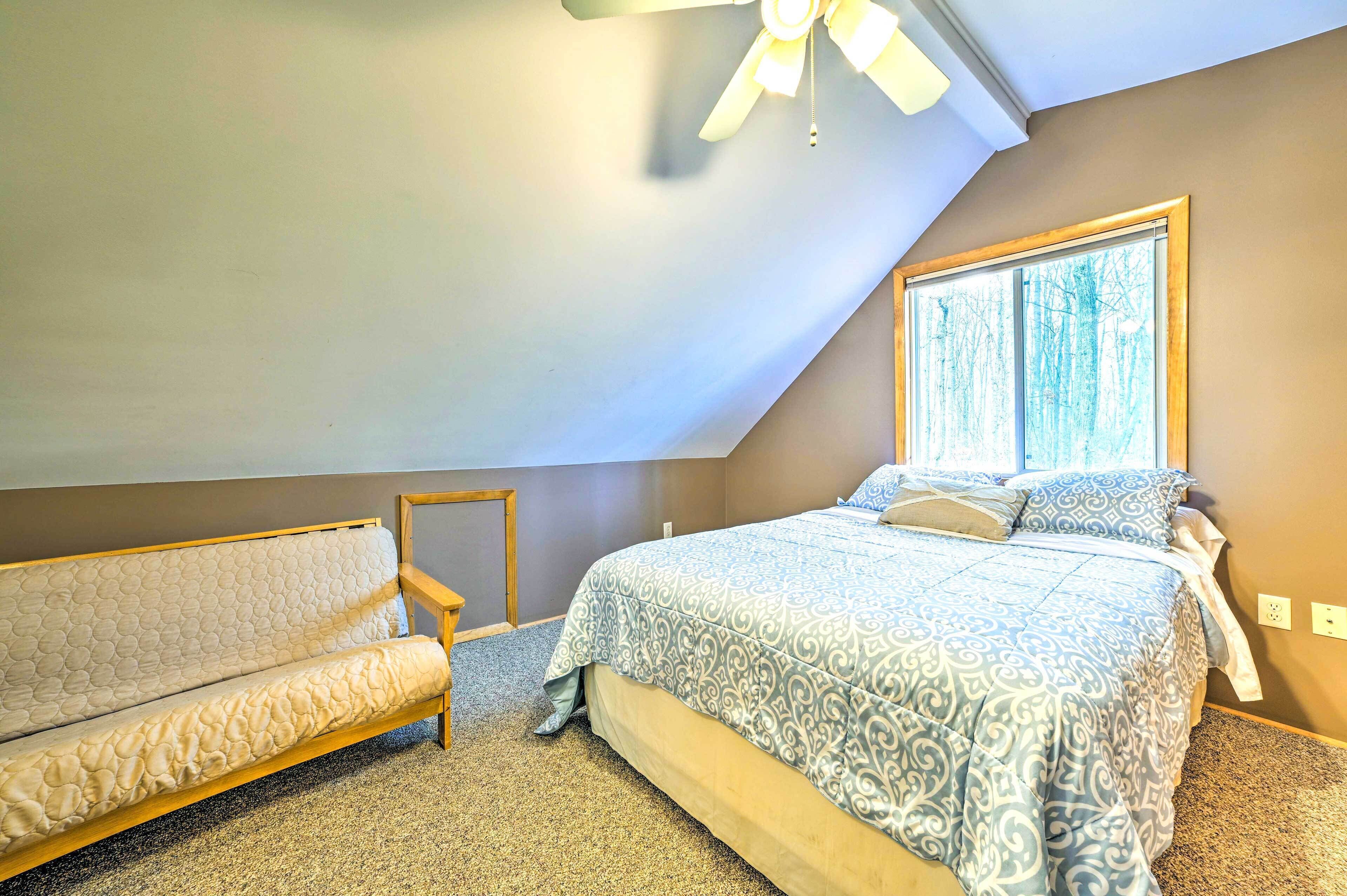 A bedroom with a bed, a sofa, a ceiling fan, and a window with a view of trees.