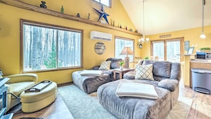 A cozy living room with large windows, a fireplace, and a kitchen area. - Higgins Lake Retreat - Hike, Swim, Relax! (Roscommon)