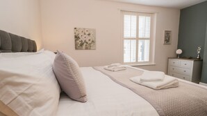 Ein ordentlich bezogenes Bett mit weißen Leinen und einem grauen Kopfteil, ein Fenster mit Jalousien, ein kleiner Nachttisch mit Spiegel und ein an der Wand hängendes Bild.