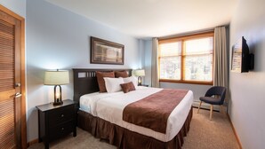 A hotel room with a large bed, a nightstand with a lamp, a chair, and a TV mounted on the wall.