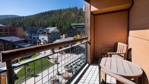 A balcony with a wooden table and chairs, overlooking a town with buildings and a forested hill.